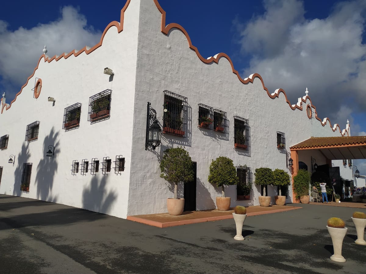 La Hacienda Valdeflores: fachada encalada con remate ondulado en almagra, rejas y faroles de forja y árboles en macetas de barro